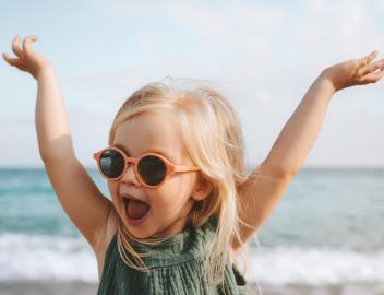 Mädchen mit Sonnenbrille am Strand streckt die Arme in die Luft.