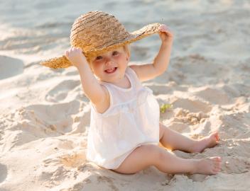 Ein kleines Mädchen im weißen Kleid mit Sonnenhut sitzt am Strand